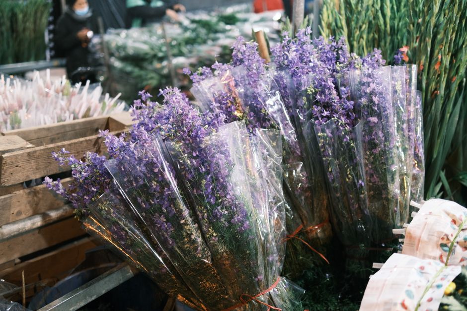 A close-up view of multiple bouquets of fresh purple lavender flowers arranged in clear cellophane wrapping with orange-brown ties, displayed outdoors on a wooden surface at a flower market. The laven