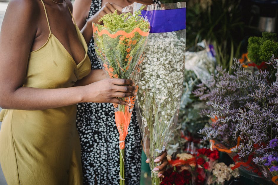 A woman wearing a light yellow sleeveless dress is holding a bouquet of green and orange flowers wrapped in clear plastic with orange and green accents. Behind her, a person in a black and white spott