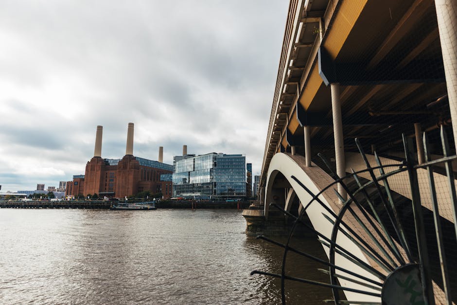A view of the River Thames in London featuring the Battersea Power Station in the background with its iconic four tall chimneys and brick facade, under an overcast sky. In the foreground, part of the 