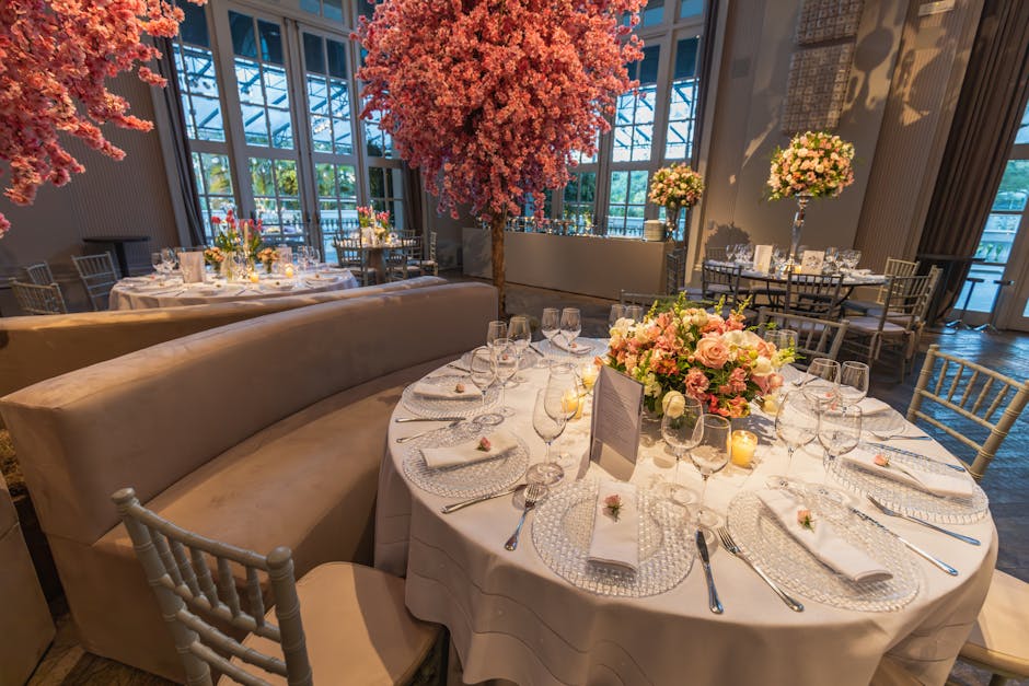 A wedding reception setup featuring a round table adorned with a large, lush floral centerpiece consisting of pink roses, cream and blush-colored flowers, and greenery, all arranged in a low, compact 