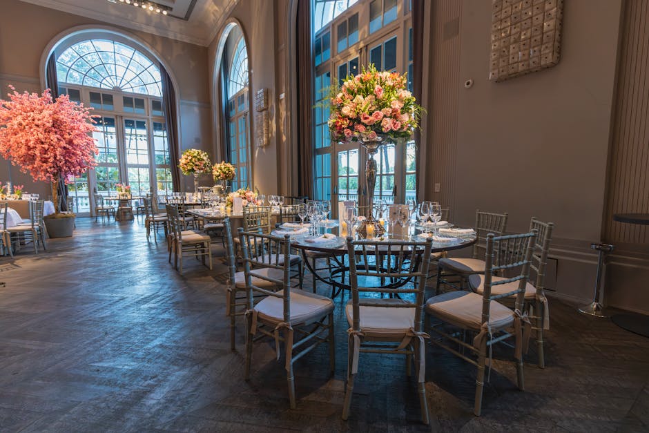 Elegant banquet room featuring tall, clear glass vases filled with large, lush arrangements of pink and white roses, hydrangeas, and greenery on round tables, with white Chiavari chairs surrounding th